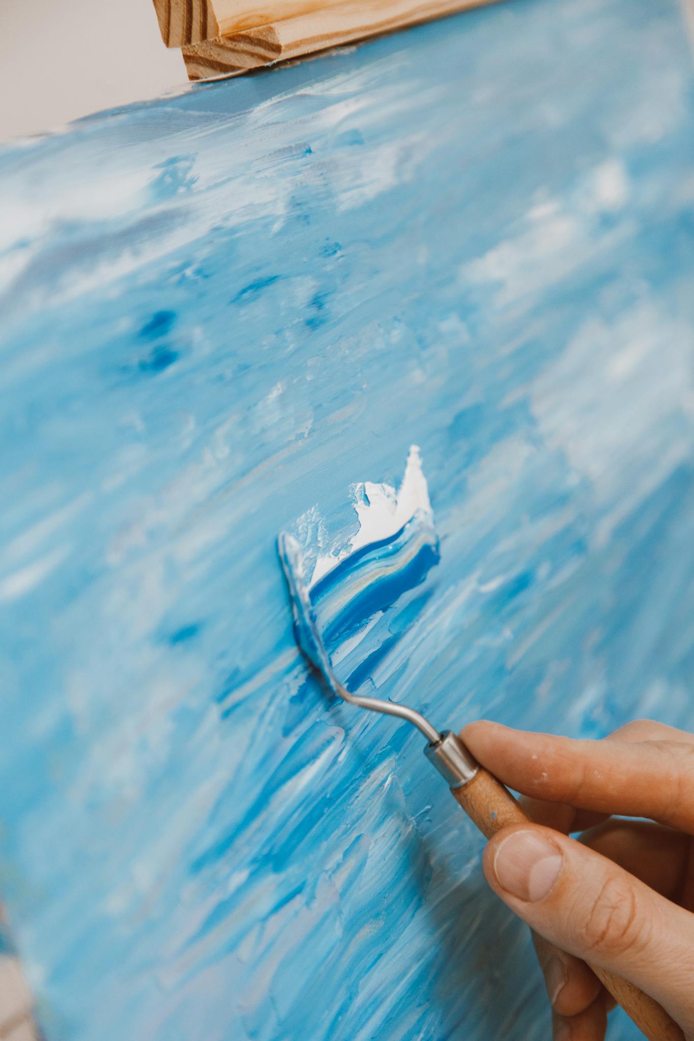 Close-up of a hand using a palette knife to create blue abstract art.